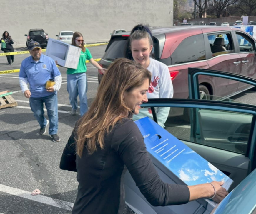 Woman loading a blue box into a car in a parking lot with other people and vehicles in the background.