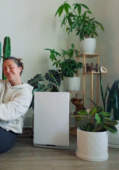 Person sitting on the floor surrounded by potted plants and an air purifier in a room with a white wall.