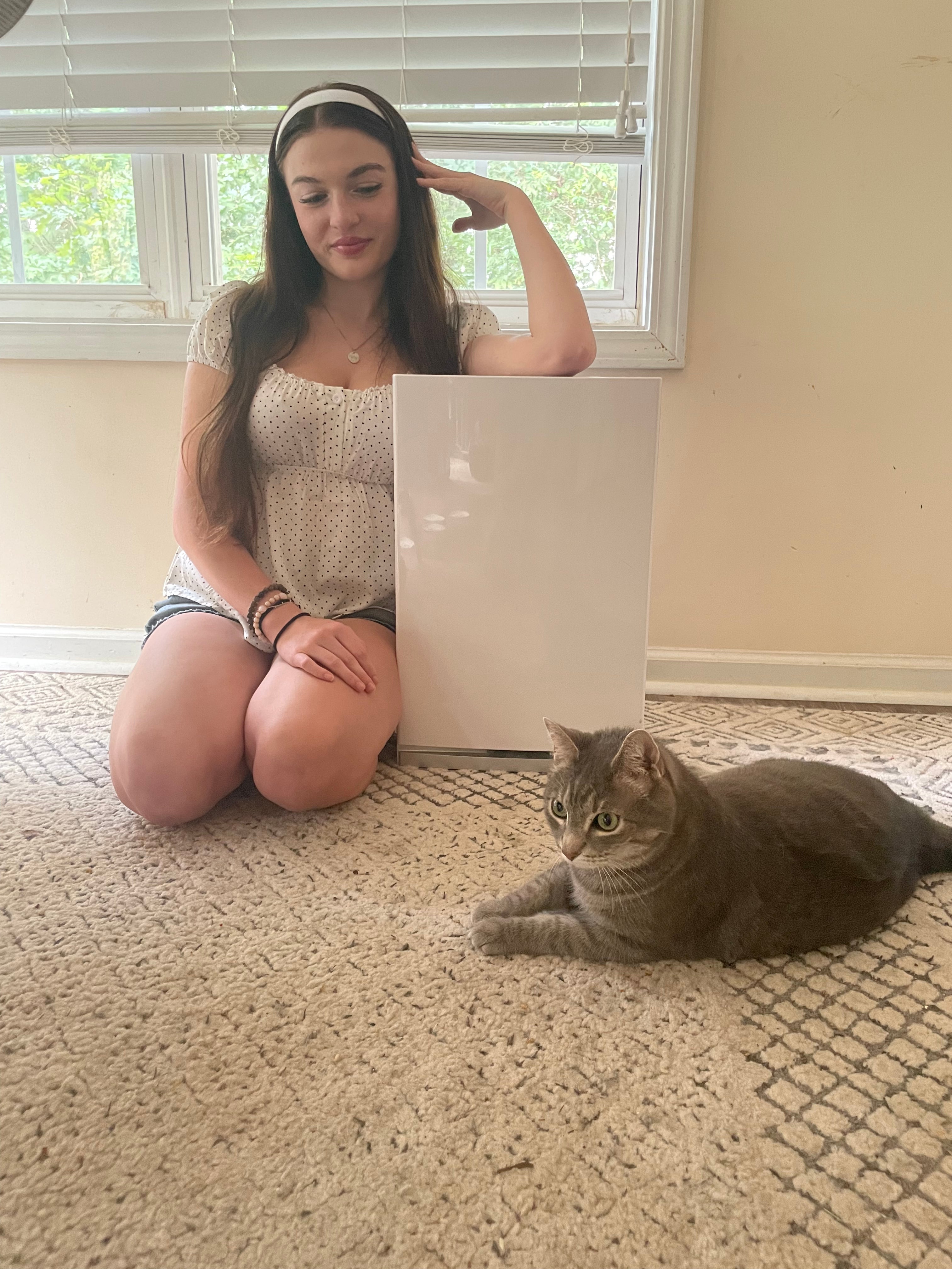 Woman sitting next to an air purifier with a cat sitting on the floor next to her in a room with carpet and window blinds.