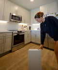 Person in a kitchen with white cabinets and stainless steel appliances, holding a white appliance.