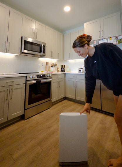 Person in a kitchen with white cabinets and stainless steel appliances, holding a white appliance.