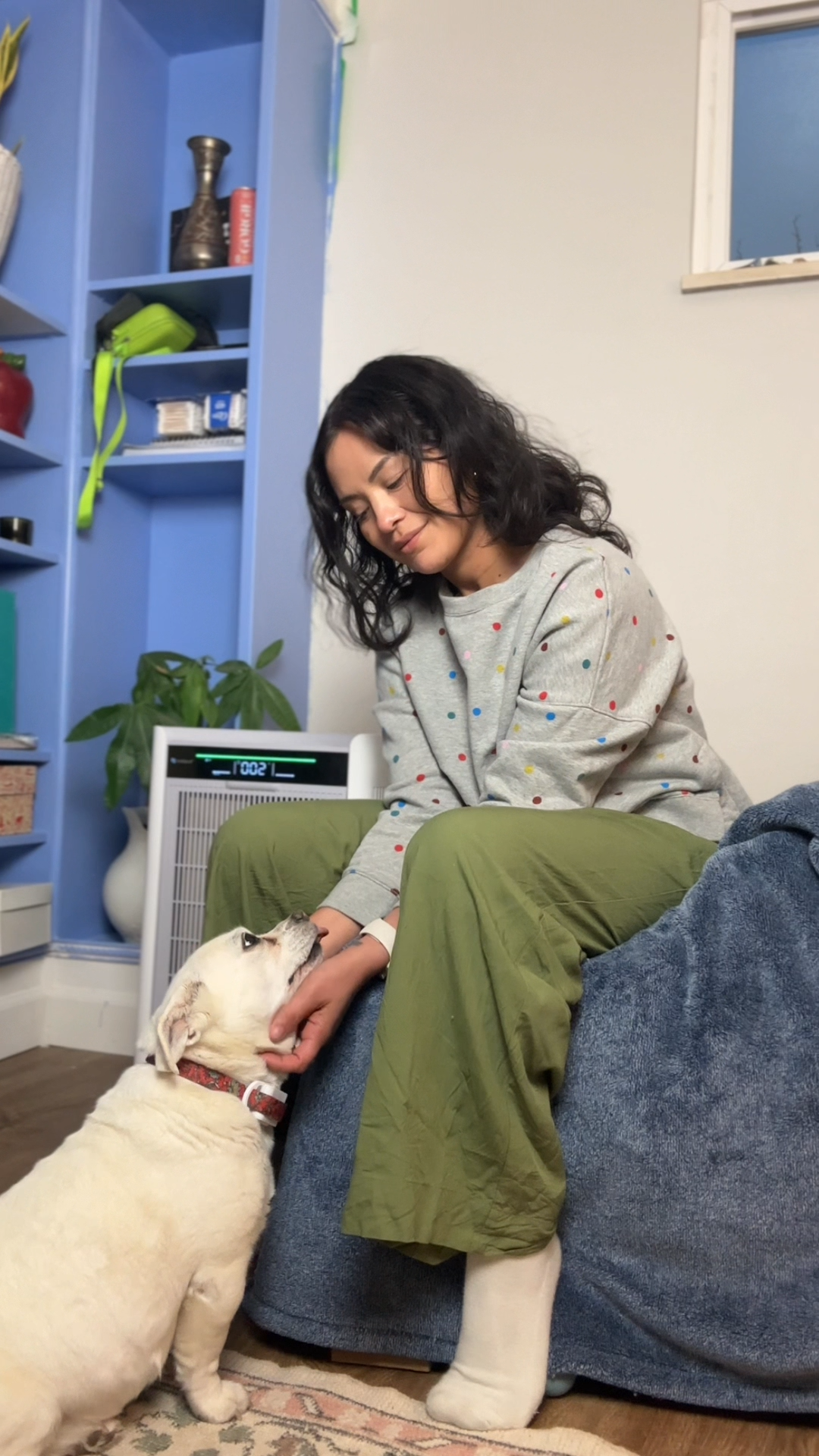 Woman sitting on a couch with a dog, next to an air purifier, in a home setting.