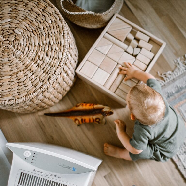 A child with blonde hair is playing with a set of wooden blocks and a toy dinosaur in a room that has wooden floors and an Intellipure air purifier.