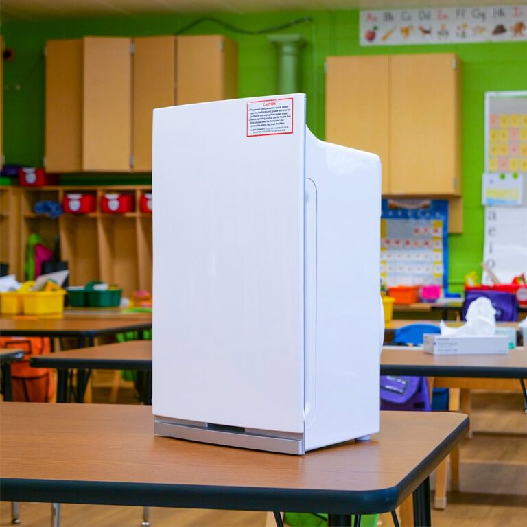 An Intellipure Compact air purifier is displayed on a wooden table in a classroom that has green walls and toy bins in the background.